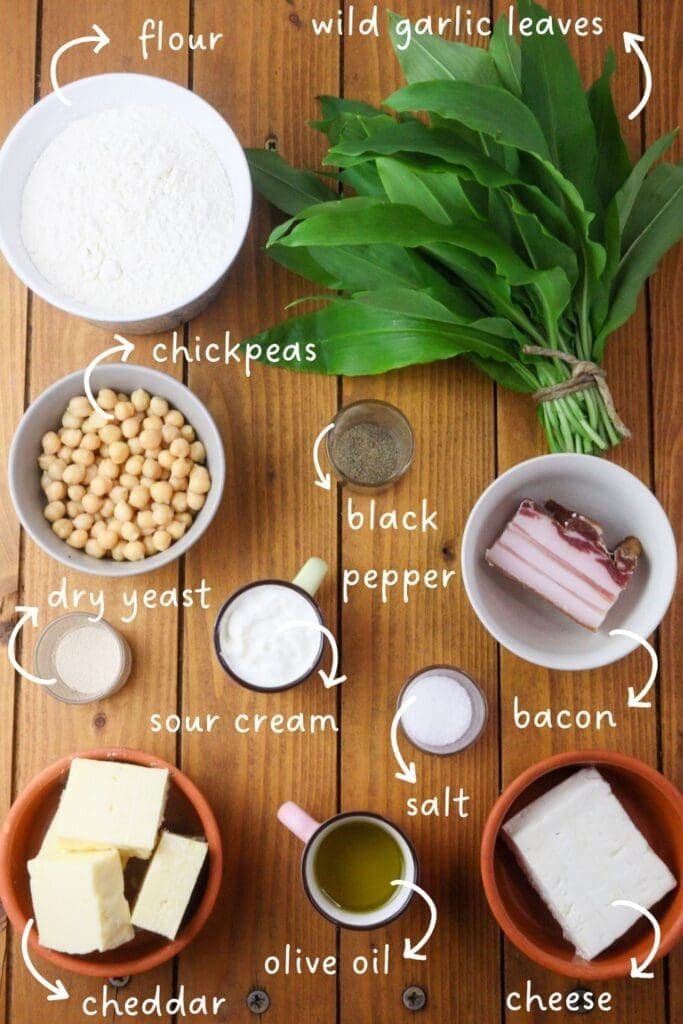 gathering ingredients for wild garlic pizza: flour, wild garlic leaves, sour cream, cheddar cheese, feta cheese, smoked bacon, cooked chickpeas, olive oil, salt and black pepper