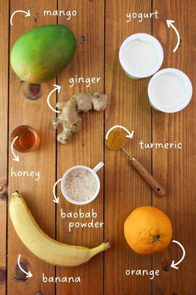 Image of ingredients needed for baobab smoothie with ginger: baobab powder, fresh ginger, yogurt, banana, orange, mango, honey and turmeric.