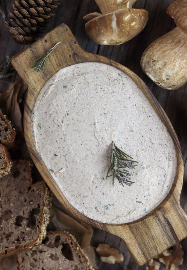 A wooden bowl of porcini mushroom walnut pate, decorated with rosemary with homemade bread and porcini mushrooms in the background.