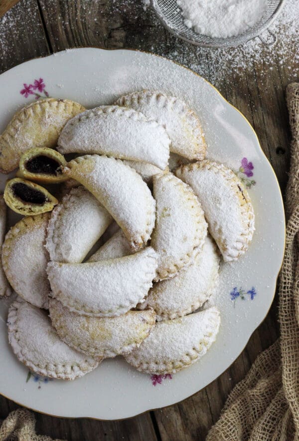 A plate of mouthwatering empanada cookies with one cookie cut in half where plum jam is visible and with a strain full of powdered sugar in the background