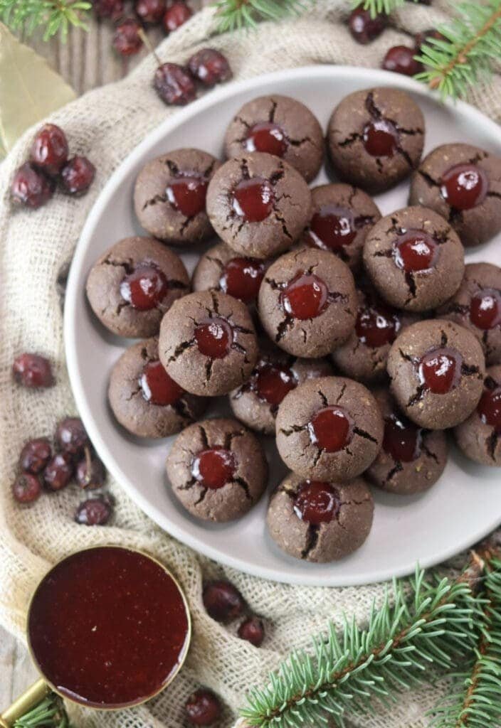 A full plate of thumbprint buckwheat cookies with jam, with a cup of strawberry jam and spruce branches in the background