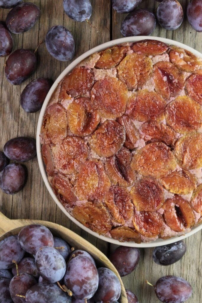 Upside down plum cake with buckwheat on a plate, surrounded with fresh plums on a wooden table