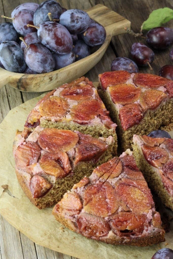 Serving slices of plum cake with buckwheat and honey by a traditional recipe, with fresh plums in the background.