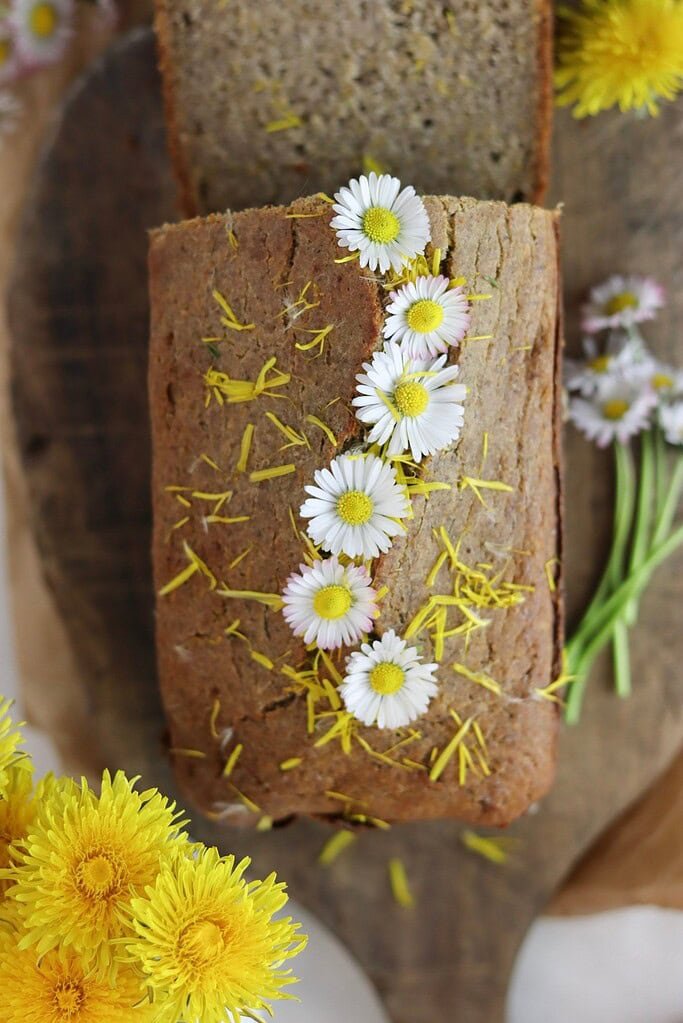 A beautiful loaf of dandelion bread with buckwheat flour and honey, with daisies on top and dandelion flowers in the background.